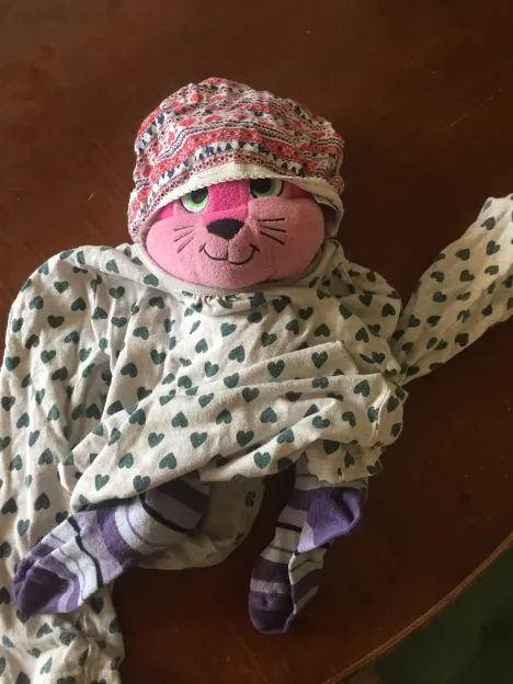 A plush cat-faced toy dressed in a patterned hat and heart-print fabric with striped socks, arranged on a wooden table in soft, warm light.